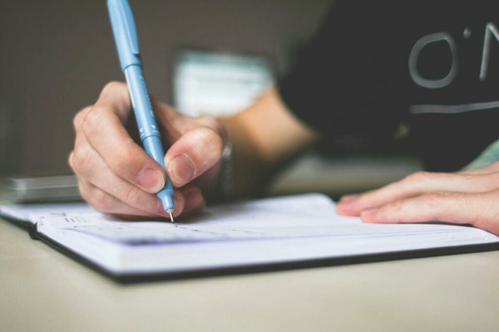 Person holding a blue ballpoint pen, writing in a notebook, symbolizing the act of practicing and refining punctuation skills.