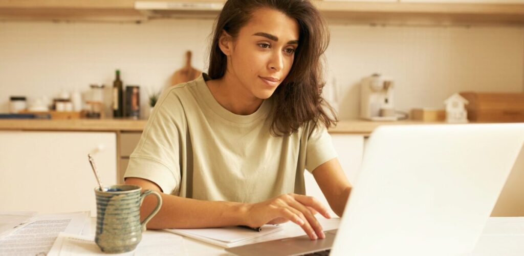 A young woman working on her laptop with papers scattered around her, symbolizing her focusing on mastering comma rules for clear, effective writing.